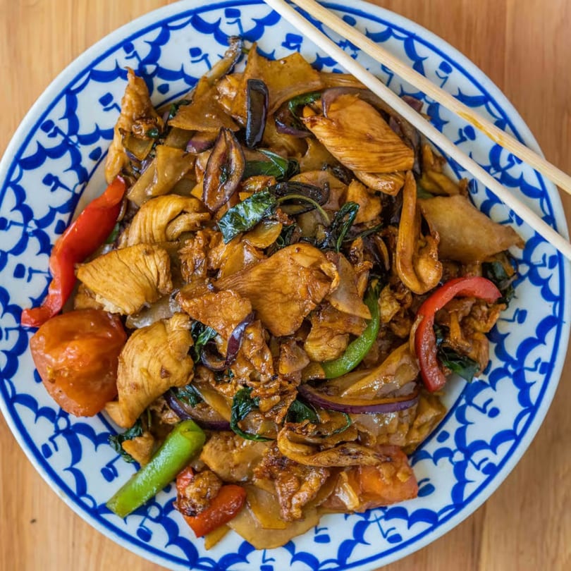Chicken Pad See Ew with wide noodles, red peppers, and green beans on a blue and white plate.