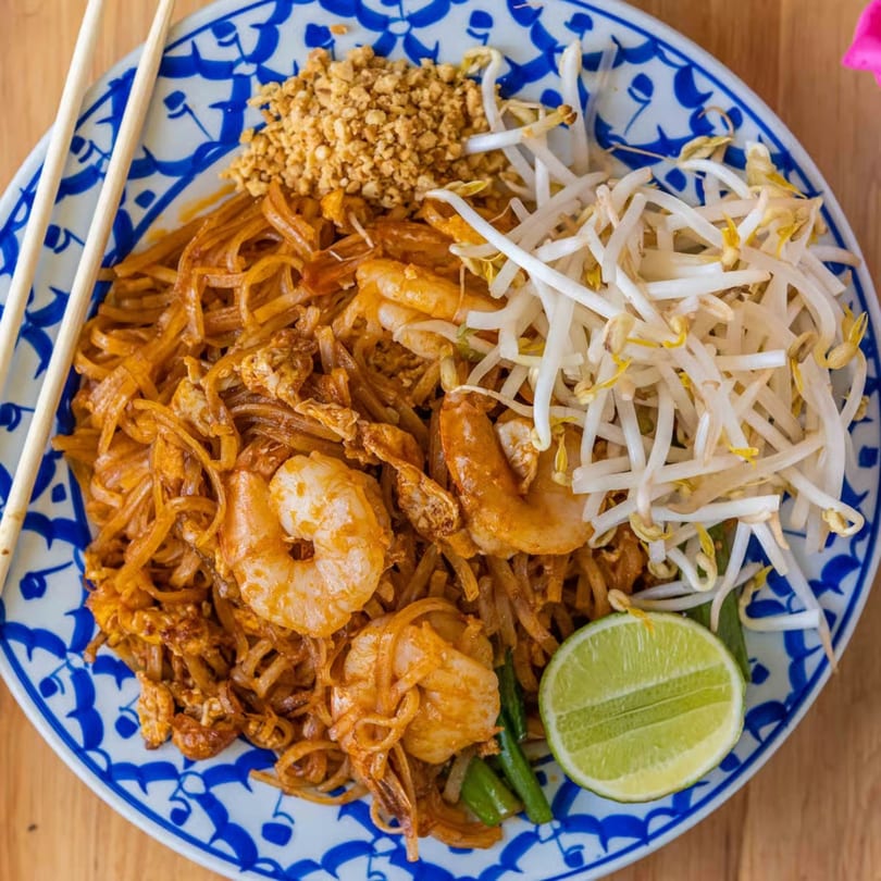 Shrimp Pad Thai with bean sprouts, crushed peanuts, and a lime wedge on a blue and white plate.